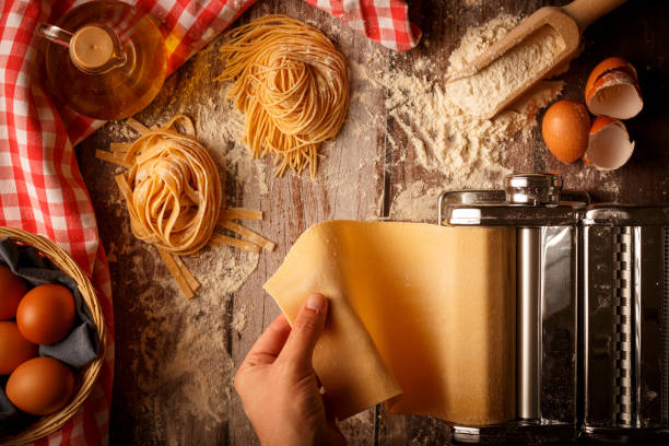Mano che usa macchina per pasta su piano infarinato, con uova, farina, olio e tagliatelle.